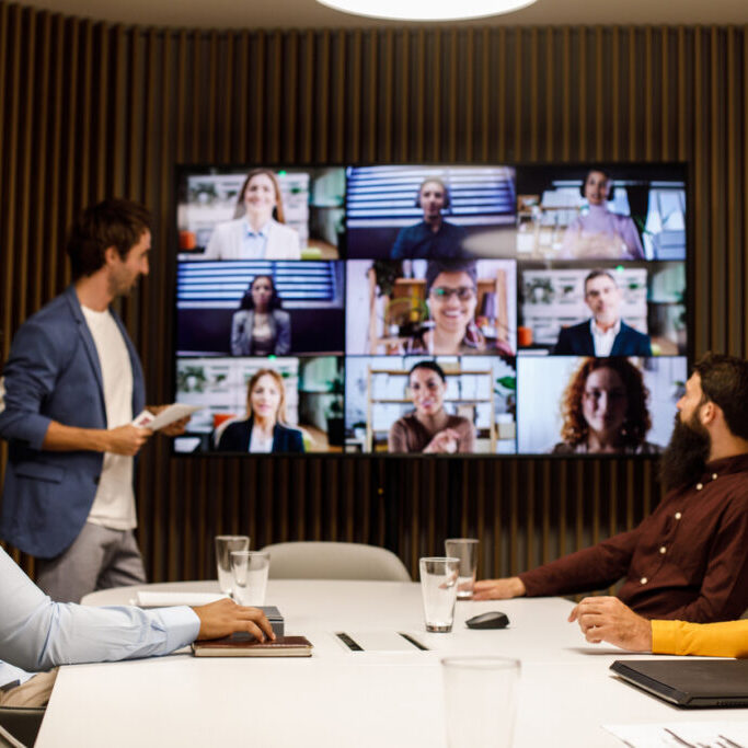 Four businessmen sitting around a conference table in a board room and communicating with their colleagues over a video call. They are discussing business projects and strategies.